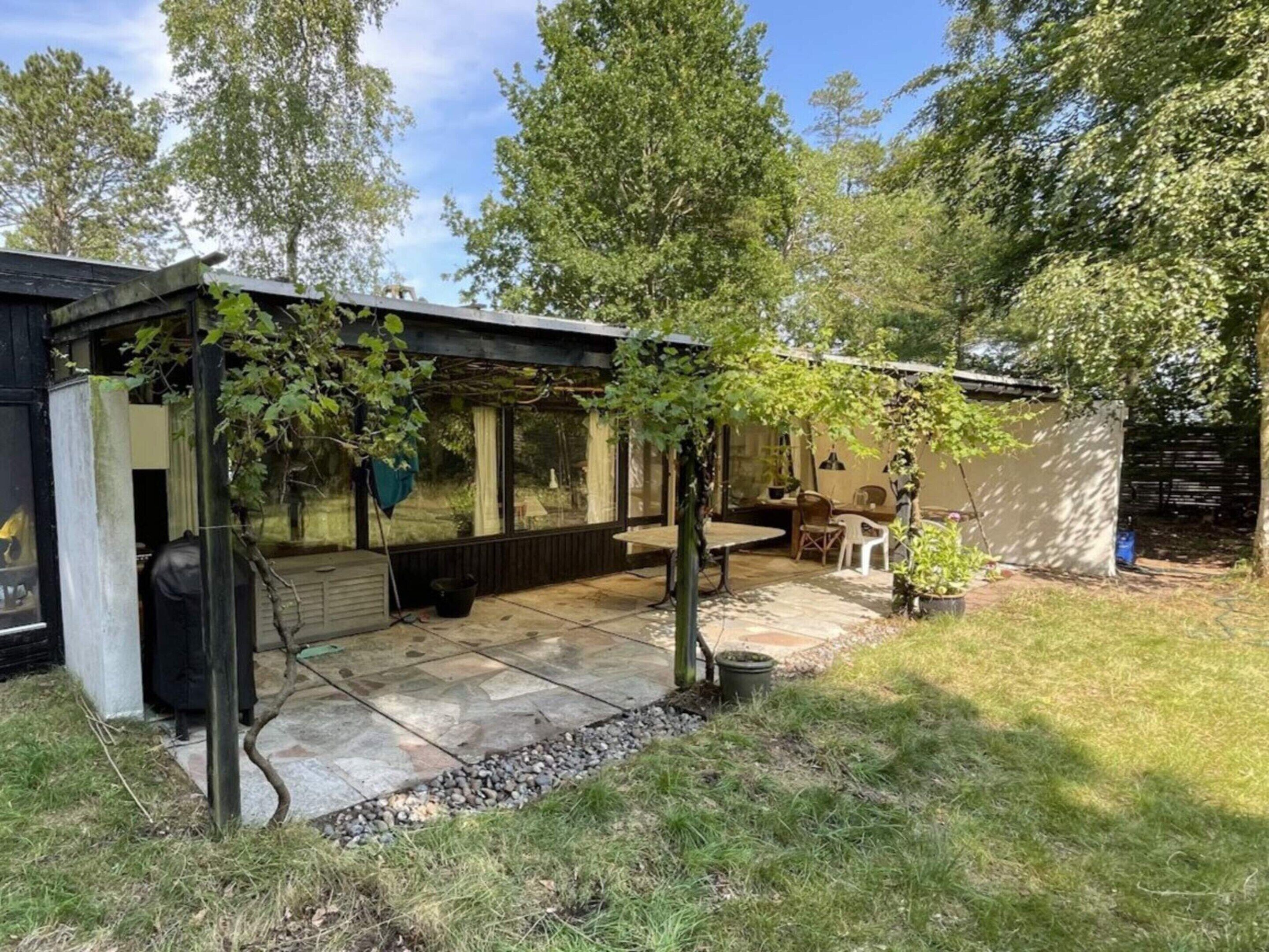 Un patio couvert avec une table et des chaises, entouré d’arbres et d’un petit abri de jardin.