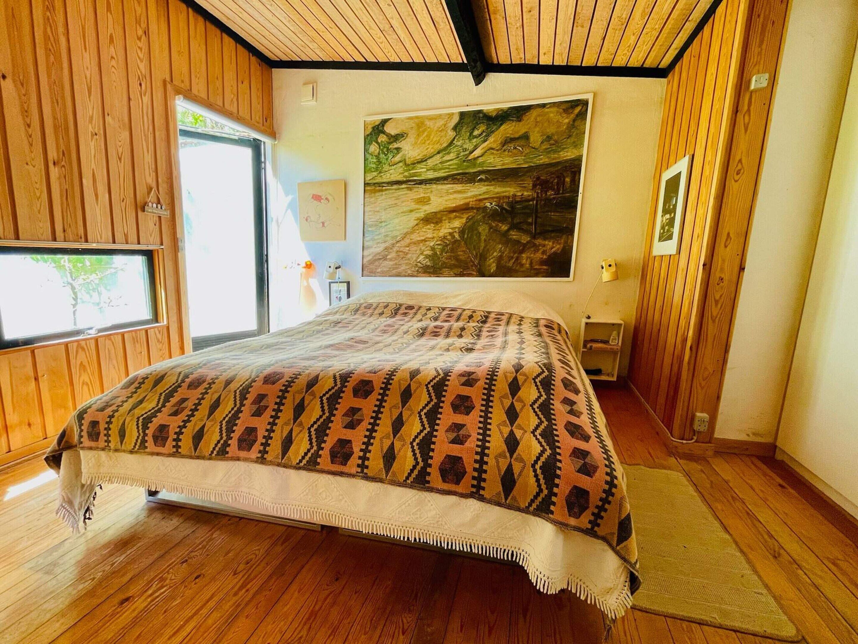 Un dormitorio con piso de madera, una cama con colcha estampada, un cuadro en la pared y una ventana con vistas.