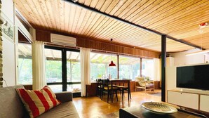 Ein Zimmer mit Holzboden, einem Sofa mit rotem und weißem Kissen, einem Esstisch mit Stühlen, einem Fernseher und großen Fenstern.
