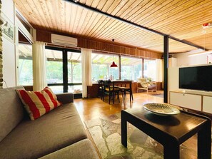 A room with a wooden ceiling, a sofa with a red and white pillow, a dining table with chairs, a television, and large windows. - 4 Star Holiday Home in Vaeggerlose (Vaeggerlose)