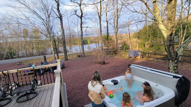 Ein Whirlpool, umgeben von Bäumen, eine Terrasse mit Stühlen und ein See im Hintergrund.