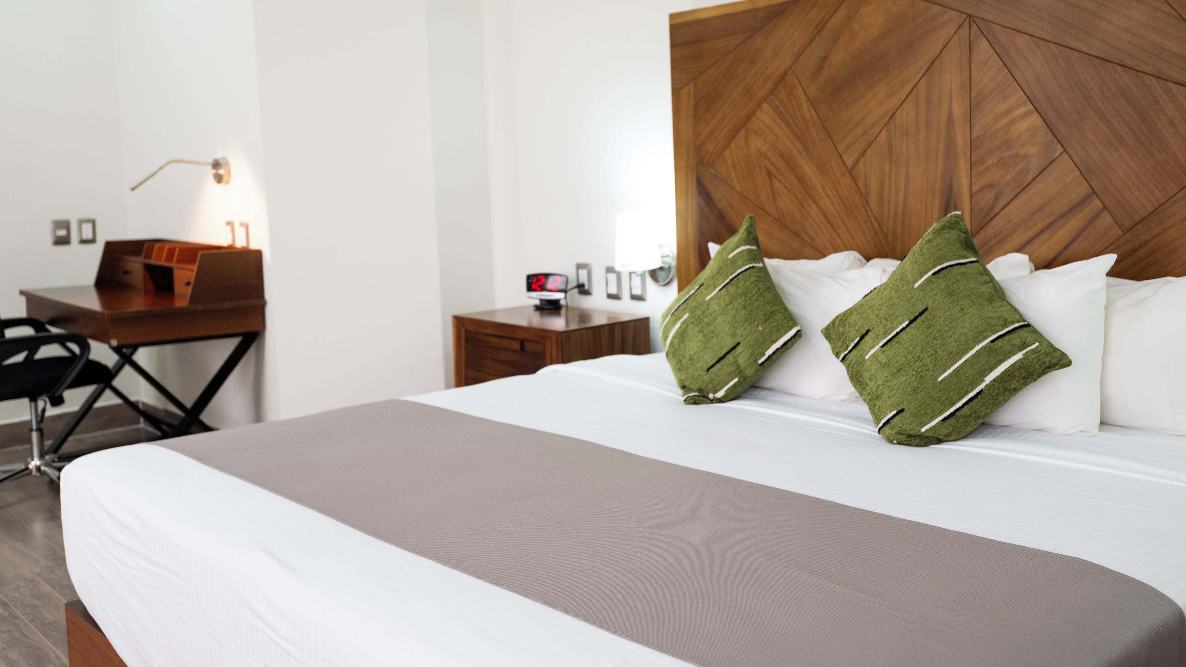 A hotel room with a large bed, a wooden headboard, a desk with a chair, a nightstand, and a wall-mounted lamp.
