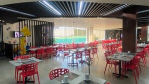 A restaurant with red chairs and tables, a high ceiling with linear lighting, and a view of greenery outside.