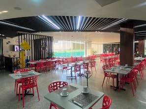 A restaurant with red chairs and tables, a high ceiling with linear lighting, and a view of greenery outside.