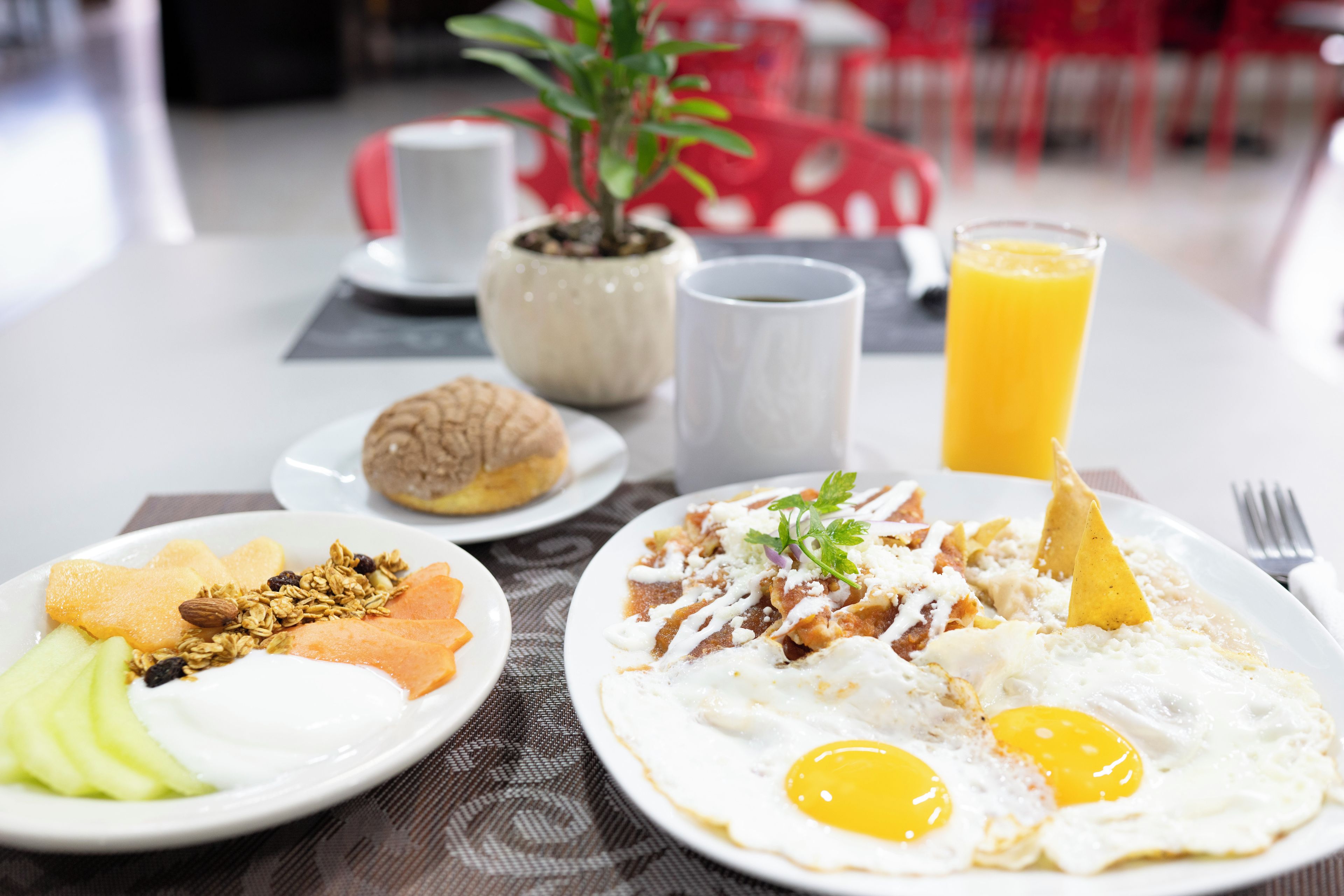 Una mesa de desayuno con platos de comida, un vaso de jugo de naranja y una planta en maceta.