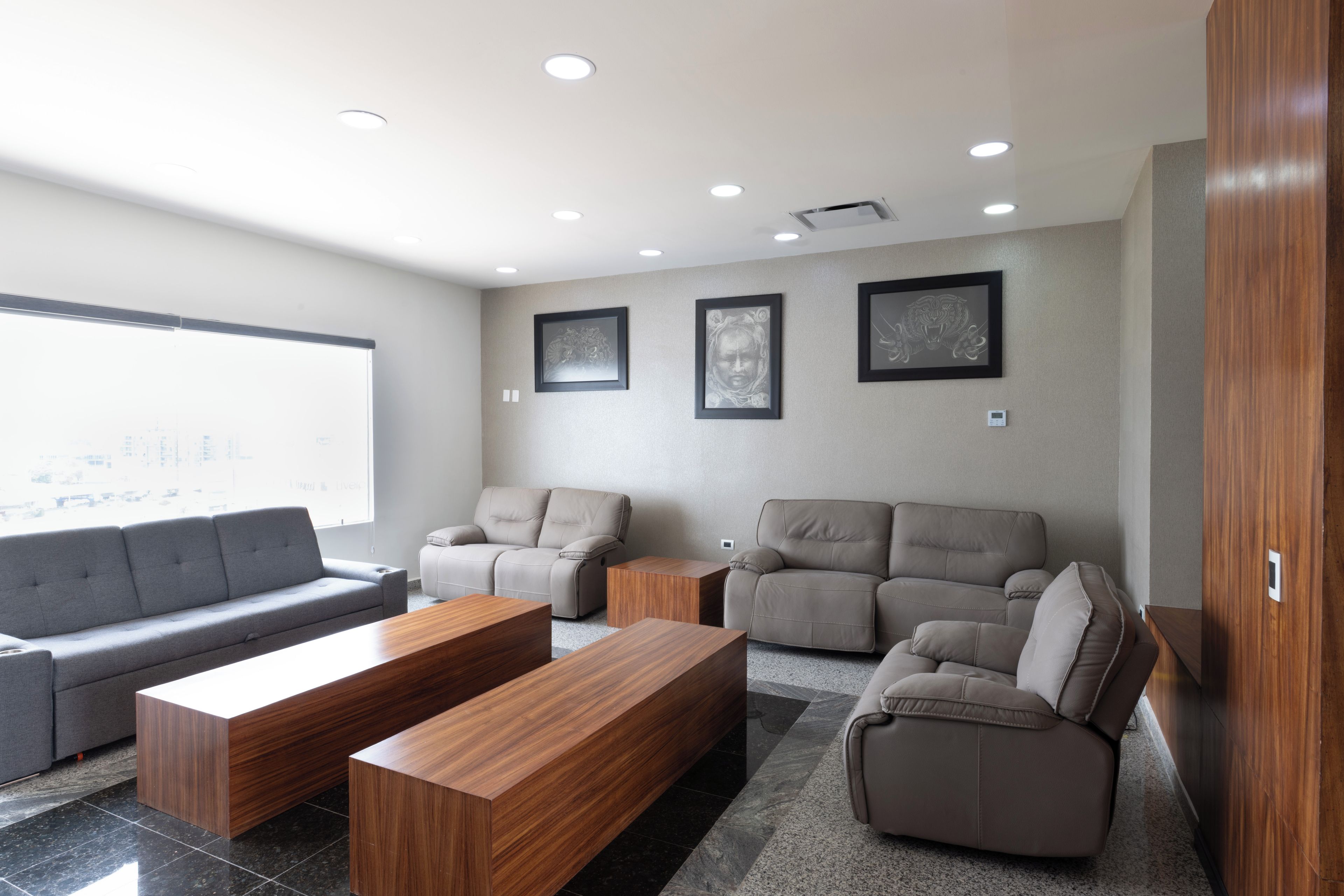 A modern living room with a grey sofa, two armchairs, and a wooden coffee table. There are three framed artworks on the wall and a large window providing a view of the city.