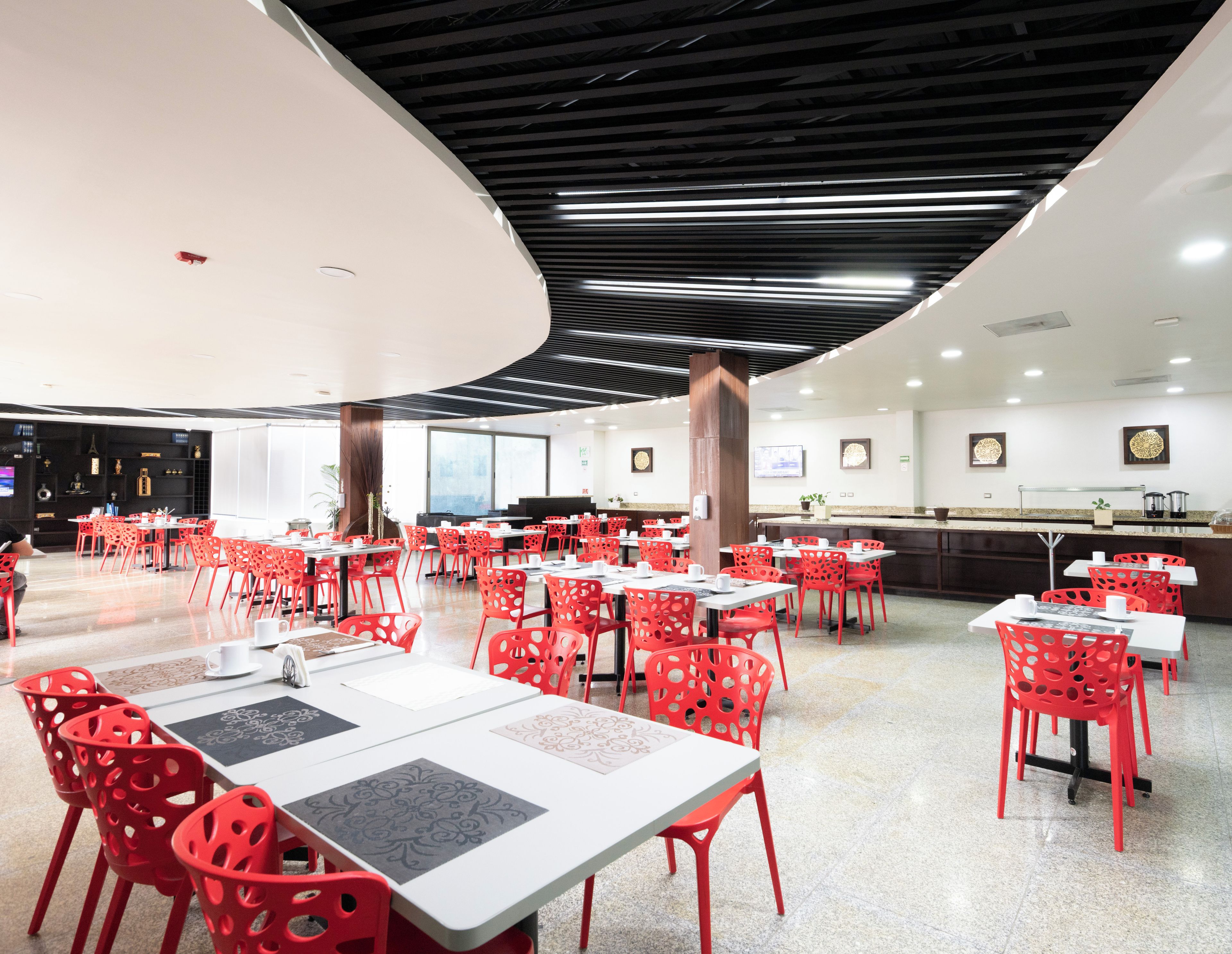 Un amplio comedor interior con mesas múltiples y sillas rojas, con un diseño de techo moderno y una barra con máquina de café.