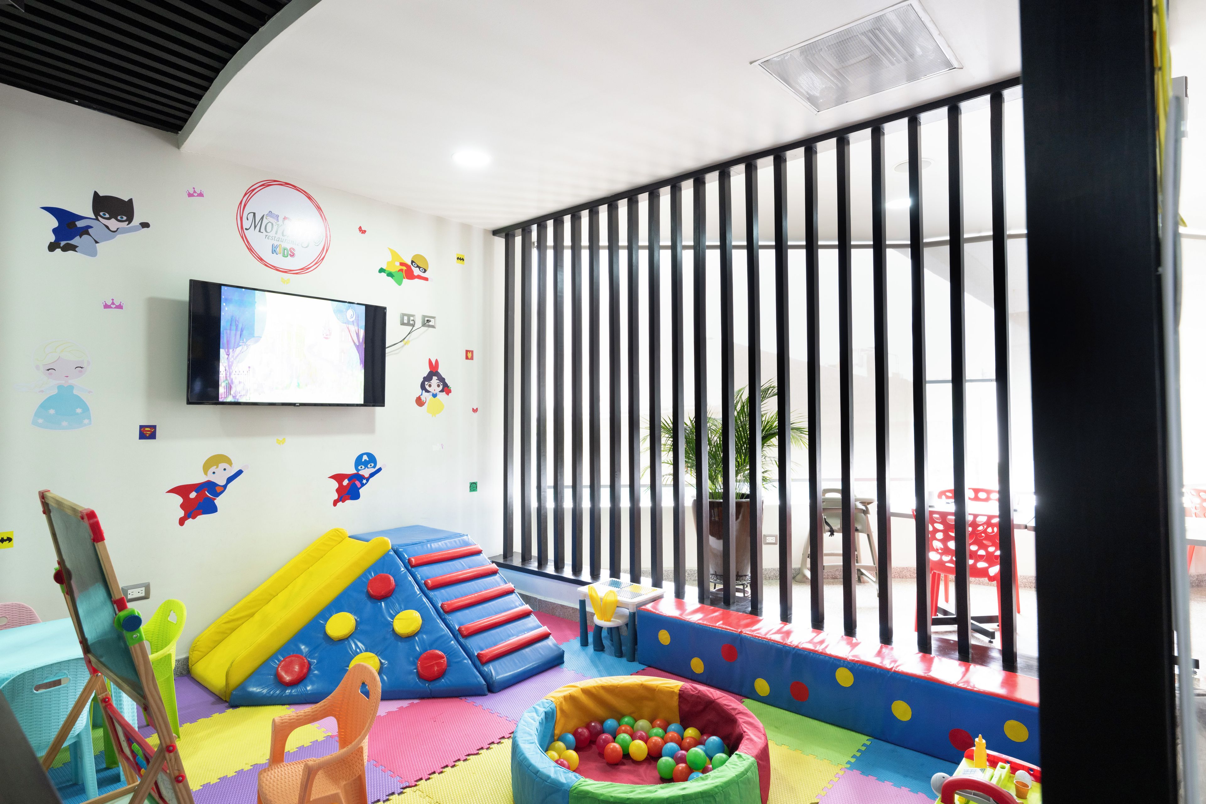 Zona de juegos para niños con tobogán, estructuras acolchadas y un televisor en la pared.