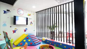 Zona de juegos para niños con tobogán, estructuras acolchadas y un televisor en la pared.