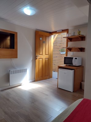 A compact room with a wooden door, a small refrigerator, a microwave, and a built-in shelf with dishes.