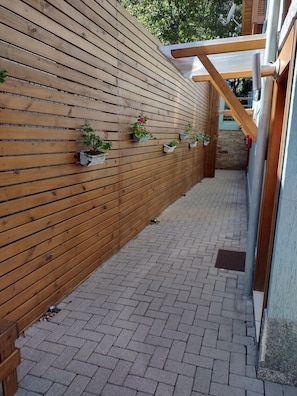 A wooden wall with potted plants, a brick pathway, and a wooden structure with a glass roof.