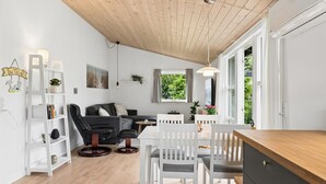 Een moderne woonkamer met een houten plafond, witte muren, een grijze bank, een eettafel met witte stoelen en een keuken met een houten aanrecht.