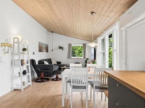 Ein modernes Wohnzimmer mit Holzboden, weißen Wänden, einem grauen Sofa, einem Esstisch mit weißen Stühlen und einer Küchenzeile mit Arbeitsplatte aus Holz.