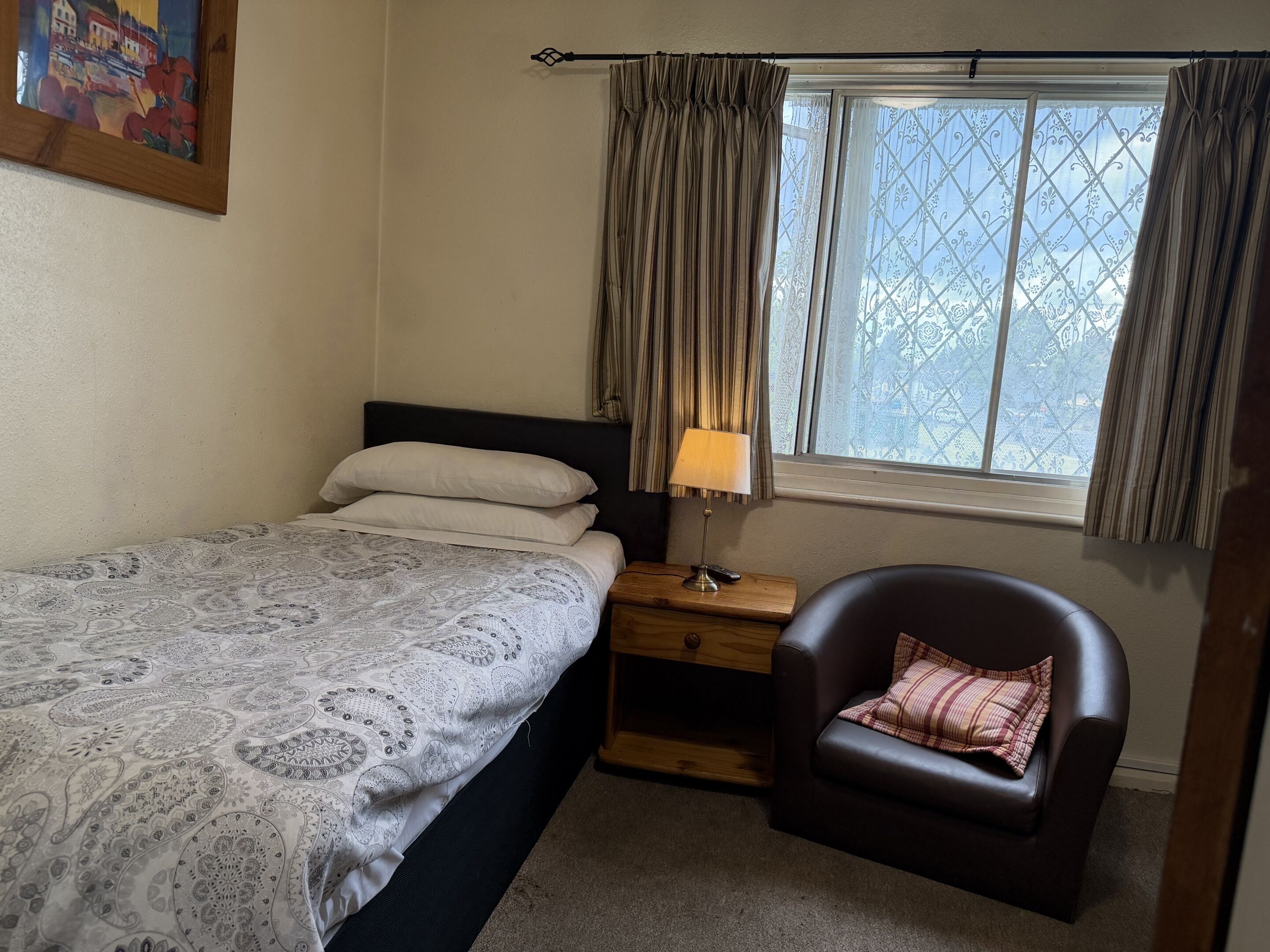 Une chambre à coucher avec un lit, une table de chevet, une chaise, une fenêtre avec des rideaux et un tableau au mur.