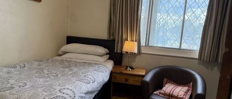 A bedroom with a bed, a nightstand, a chair, a window with curtains, and a painting on the wall.