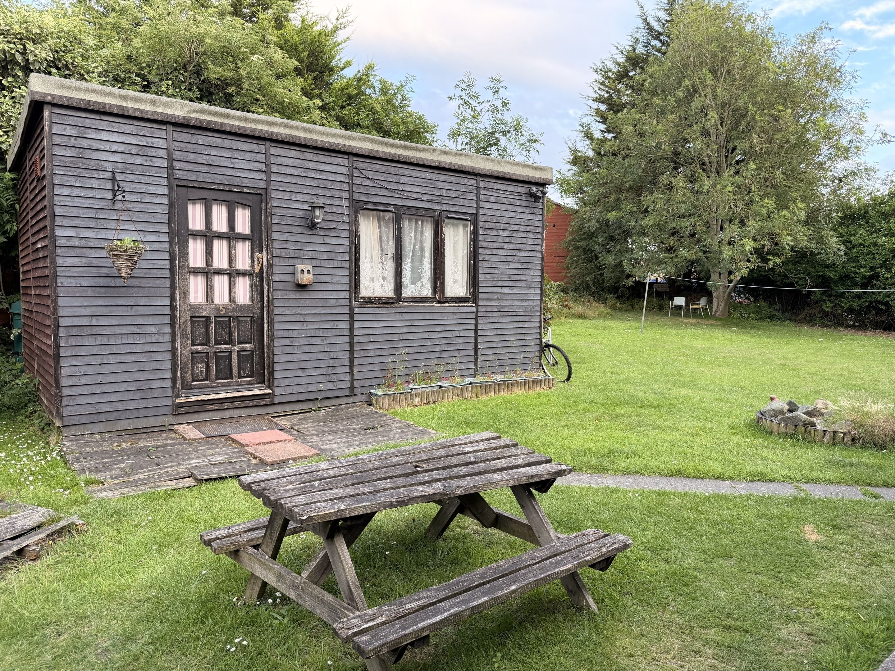 A small wooden cabin with a door, windows, and a picnic table in a grassy yard.