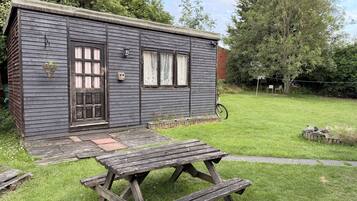 A small wooden cabin with a door, windows, and a picnic table in a grassy yard.