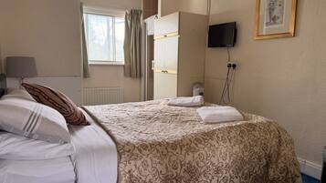 A hotel room with a bed, a bedside table with a lamp, a window with curtains, a wall-mounted TV, and a framed picture.