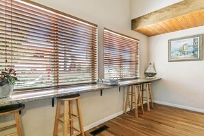 A kitchen with two windows, wooden stools, a countertop, and a painting on the wall.