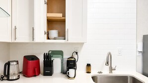 Una cucina con mobili bianchi, un lavello in acciaio inossidabile, una caffettiera rossa e un portachiodi.