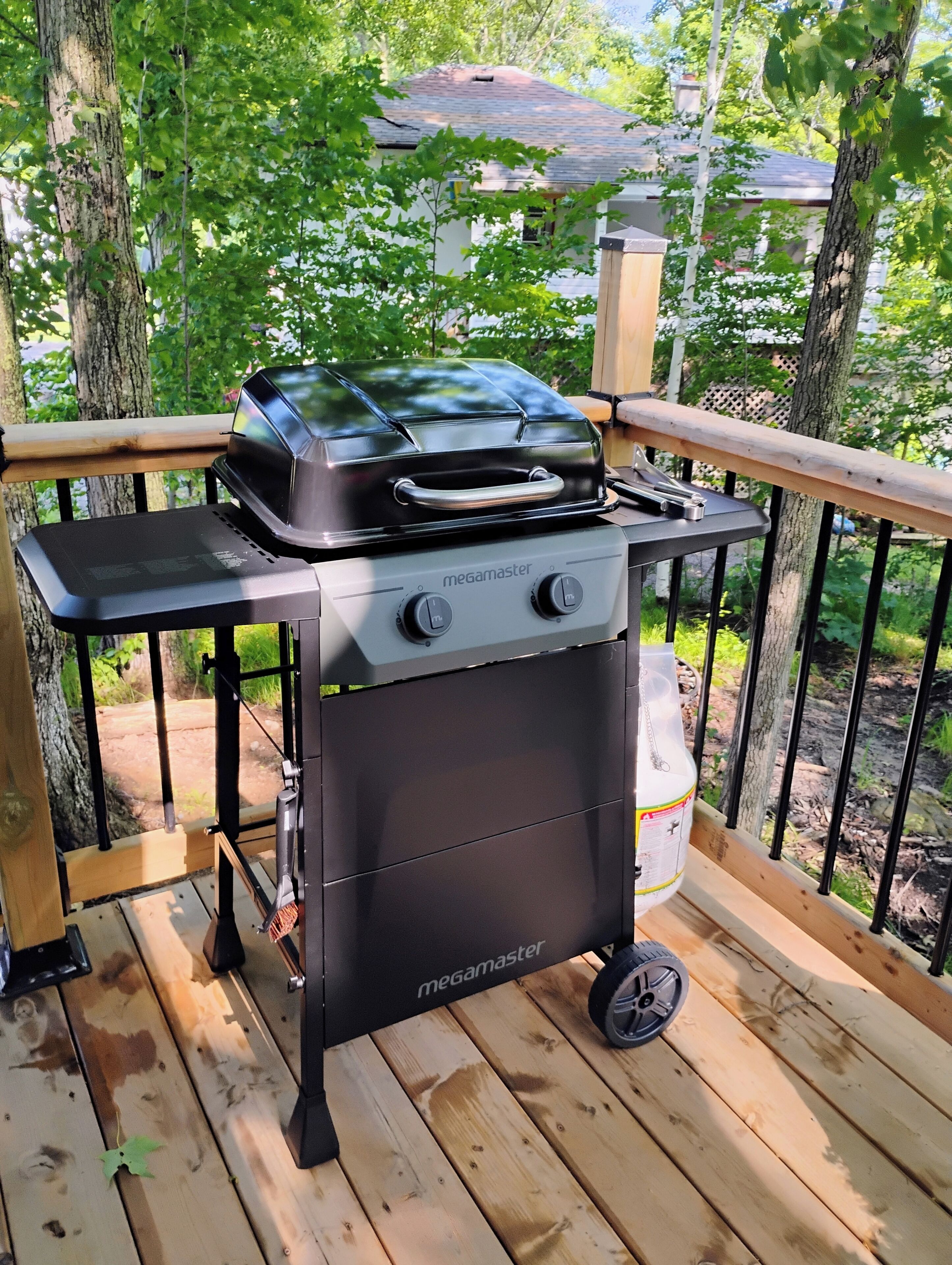 A portable Megamaster grill on a wooden deck with a railing, surrounded by trees.