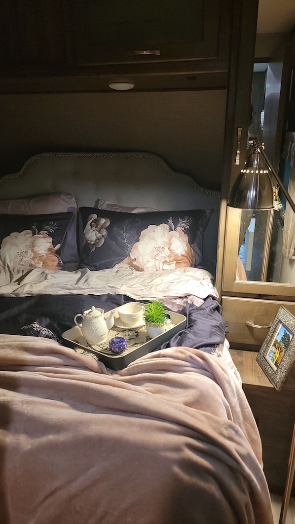 A bed with a floral duvet, a bedside table with a lamp, and a tray with a teapot and cups.