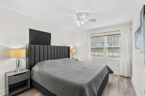 Een slaapkamer met een bed, nachtkastjes, een plafondventilator, een raam met gordijnen en een kunstwerk aan de muur.