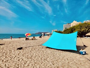 Ein Strand mit einer blauen Markise, einem Strandkorb und einer Kühlbox im Sand.