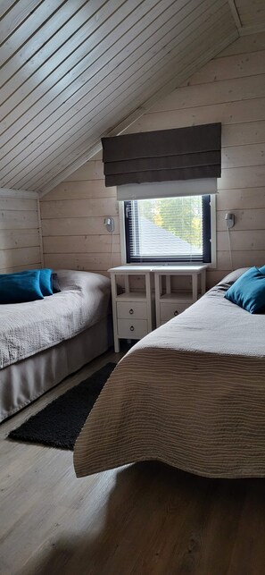 A room with two beds, a small table, a window with blinds, and a ceiling with wooden beams.