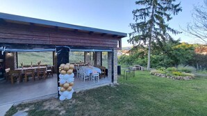 Um pátio externo com mesas e cadeiras, um arco de balões decorativo e vista para uma área gramada com árvores.