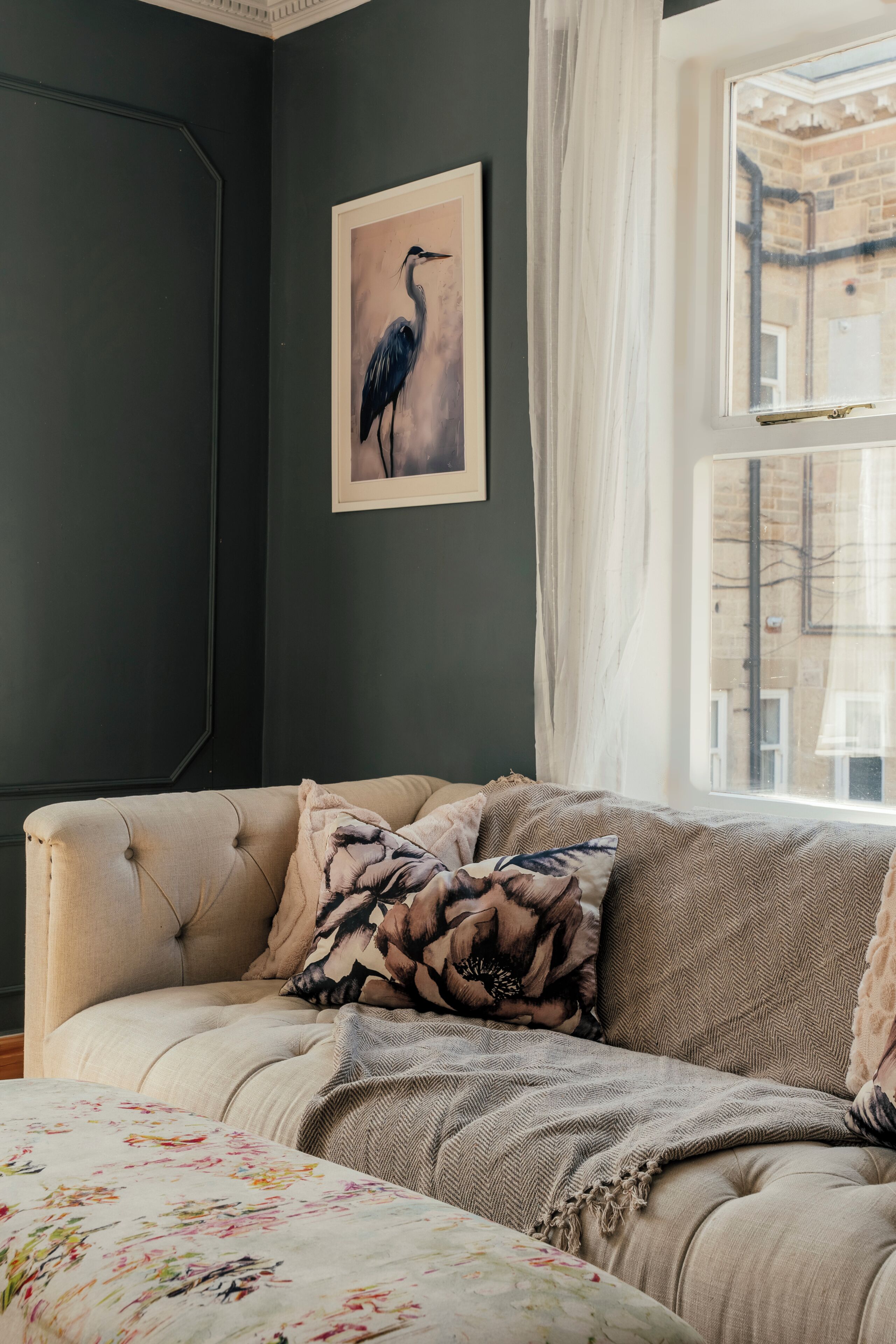 A room with a tufted sofa, a framed bird painting, and a window with a view of buildings.