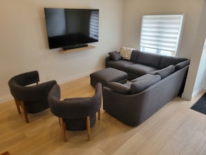 Een woonkamer met een flatscreen televisie aan de muur, een donkergrijze bank, twee fauteuils en een raam met zonwering.