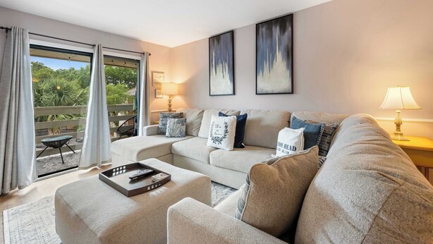 A modern living room with a sofa set, a coffee table, and a side table with a lamp. There is a balcony with curtains and a view of greenery. - Relaxing Beach Retreat Villa, Walk to Beach and Resort (Hilton Head)