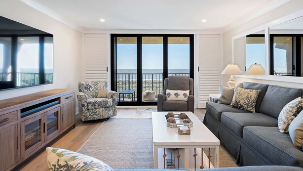 A living room with a large TV, a sofa, a chair, a coffee table, and a view through the glass doors. - Luxury Direct Oceanfront 3BR Panoramic Ocean Views (Hilton Head Island)