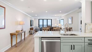 A modern kitchen with a dishwasher, a dining area with a table and chairs, and a large window offering a view. - Luxury Direct Oceanfront 3BR Panoramic Ocean Views (Hilton Head Island)