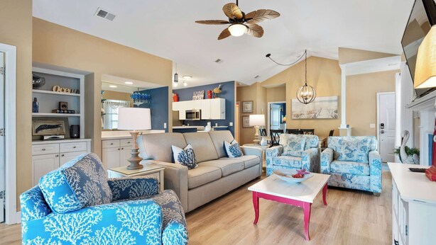 A living room with a sofa, armchairs, a coffee table, a ceiling fan, and a kitchen area with cabinets and appliances. - Beautiful & Spacious Golf View Villa Next to Pool (Hilton Head)