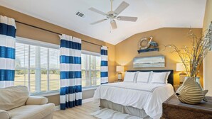 A bedroom with a bed, a ceiling fan, a window with curtains, a nightstand, a lamp, and a decorative fish on a wooden table. - Beautiful & Spacious Golf View Villa Next to Pool (Hilton Head)