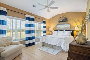 A bedroom with a bed, a ceiling fan, a window with curtains, a nightstand, a lamp, and a decorative fish on a wooden table. - Beautiful & Spacious Golf View Villa Next to Pool (Hilton Head)