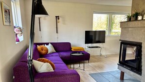 Olohuoneessa on violetti sohva, televisio, takka ja ikkuna, josta avautuu näkymä puita kohti.