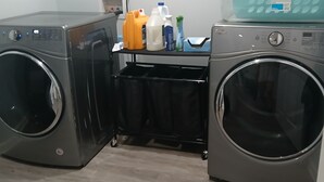 Uma área de lavanderia com duas máquinas de lavar de abertura frontal, um carrinho de armazenamento preto e uma cesta de plástico azul em cima da máquina à direita.