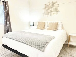 Un dormitorio con cama, mesita de noche, una obra de arte en la pared y una ventana con cortinas.