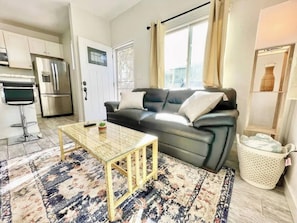 Ein Wohnzimmer mit einer Couch, einem Couchtisch, einem gemusterten Teppich, einer Küchenzeile mit Kühlschrank und einem Fenster mit Vorhängen.