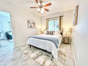 Ein Schlafzimmer mit einem Bett, einem Deckenventilator, einem Fenster mit Vorhängen, einem Nachttisch mit Lampe und einer Tür zu einem anderen Raum.
