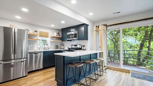 A modern kitchen with a central island, stainless steel appliances, and a large window with a view of trees. - Verdant Falls! Hot Tub & Mtn Views near Broadmoor (Colorado Springs)