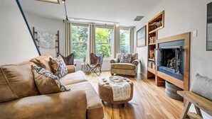 A living room with a sofa, ottoman, chairs, a fireplace, and large windows. - Verdant Falls! Hot Tub & Mtn Views near Broadmoor (Colorado Springs)