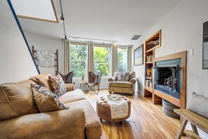 Ein Wohnzimmer mit Sofa, Hocker, Sesseln, Kamin und großen Fenstern.