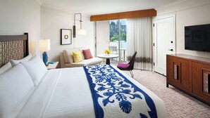 A hotel room with a large bed, a sofa, a small table with flowers, a chair, a TV, and a balcony with a view.