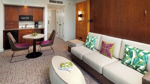 A modern hotel room with a kitchenette, a sofa set, a round coffee table, and a patterned carpet.