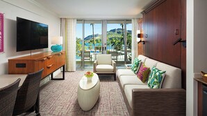 A hotel room with a flat-screen TV, a sofa, a dining table with chairs, a coffee table, and a view of the ocean and mountains.