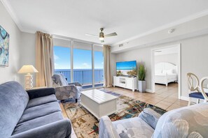 Ein Wohnzimmer mit einer Couch, einem Sessel, einem Couchtisch und einem Fernseher. Ein Balkon bietet Blick auf das Meer.